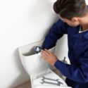 A plumber in a blue uniform repairs a toilet with tools in a bathroom, representing Bud's Plumbing & Repair Service.