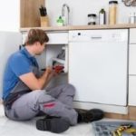 Technician repairing a dishwasher in a kitchen, focusing on appliance maintenance for Bud's Plumbing & Repair Service.