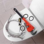 Plumbing tools on a toilet, including an adjustable wrench and hose, from Bud's Plumbing & Repair Service.