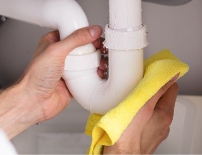 Hands inspecting a leaking plumbing joint under a sink, using a yellow cloth to catch water, illustrating the importance of detecting and repairing leaks to prevent water damage.