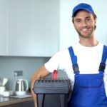 Smiling maintenance worker in blue overalls with a black toolbox in a modern kitchen, ready for plumbing services.