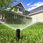 Sprinkler system watering a lush lawn outside a home, showcasing property care by Bud's Plumbing Service.