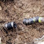 Damaged plumbing pipe leaking into muddy ground with pipe fittings, highlighting infrastructure issues with Bud's Plumbing.